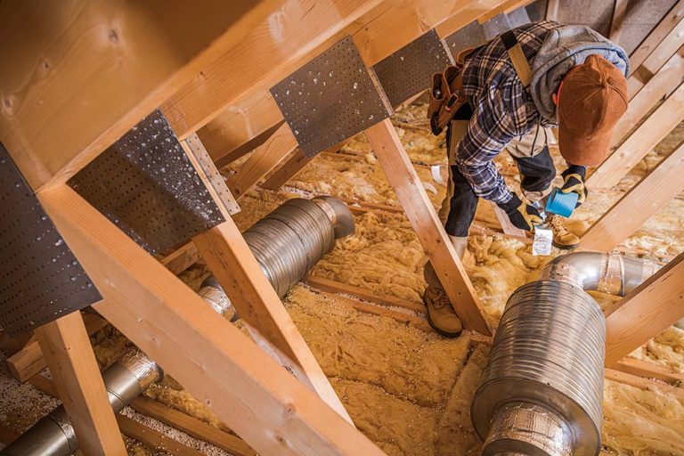 Worker applying aluminum foil tape to seal metal air ventilation ducts inside a wood-framed attic space.